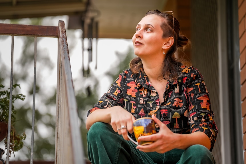 A woman with a colourful shirt holding a cup of tea while sitting on a staircase outside