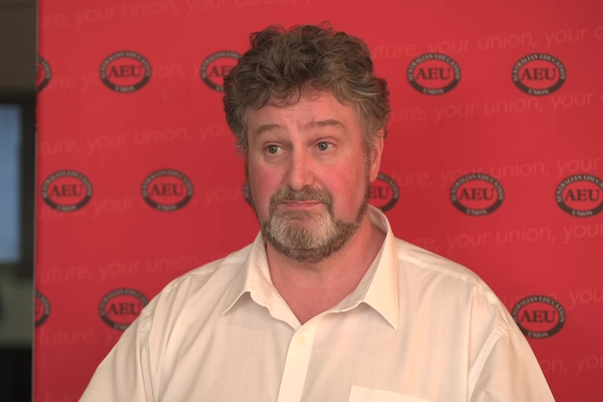 A man with curly light brown hair and beard in a cream collared shirt stands in front of a red AEU backdrop.