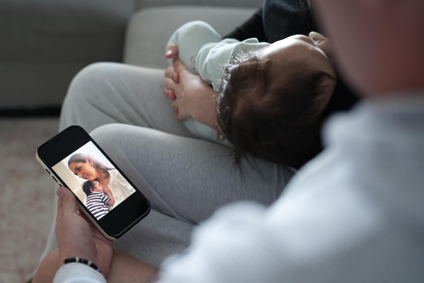 A person sits on a couch, holding a phone showing an image of a mother with a baby.
