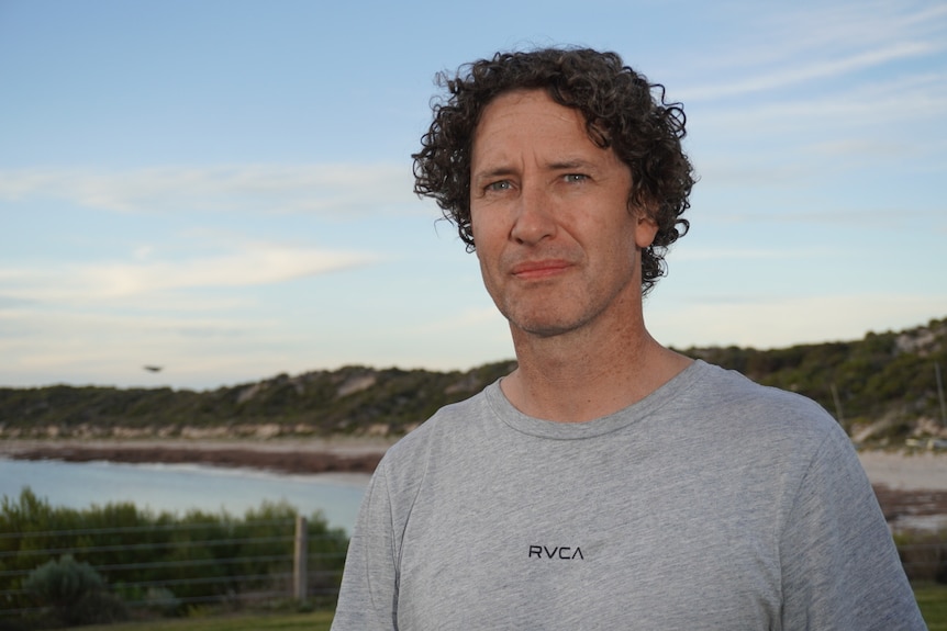 A man wearing a t-shirt stands in front of a beach