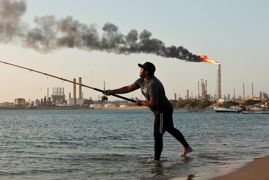 A man fishes in front of an oil refinery tower blowing flames and smoke.