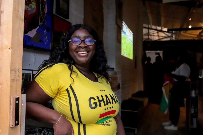 A woman wearing a yellow t-shirt with GHANA written on the front stands with a large tv screen and crowd of people behind.