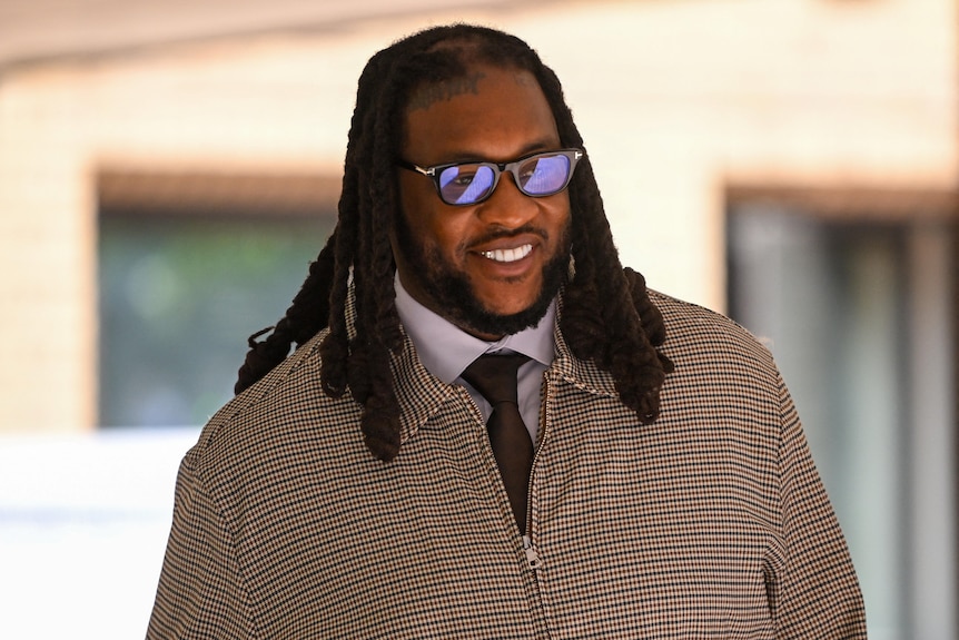A man with black long hair, a brown jacket, white shirt and brown tie wearing sunglasses.