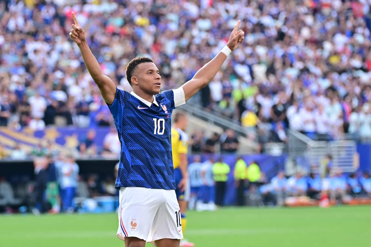 Kylian Mbappe, a French football player, dressed in blue and white with the number 10 on his chest, stands on a football pitch with his arms raised in celebration.
