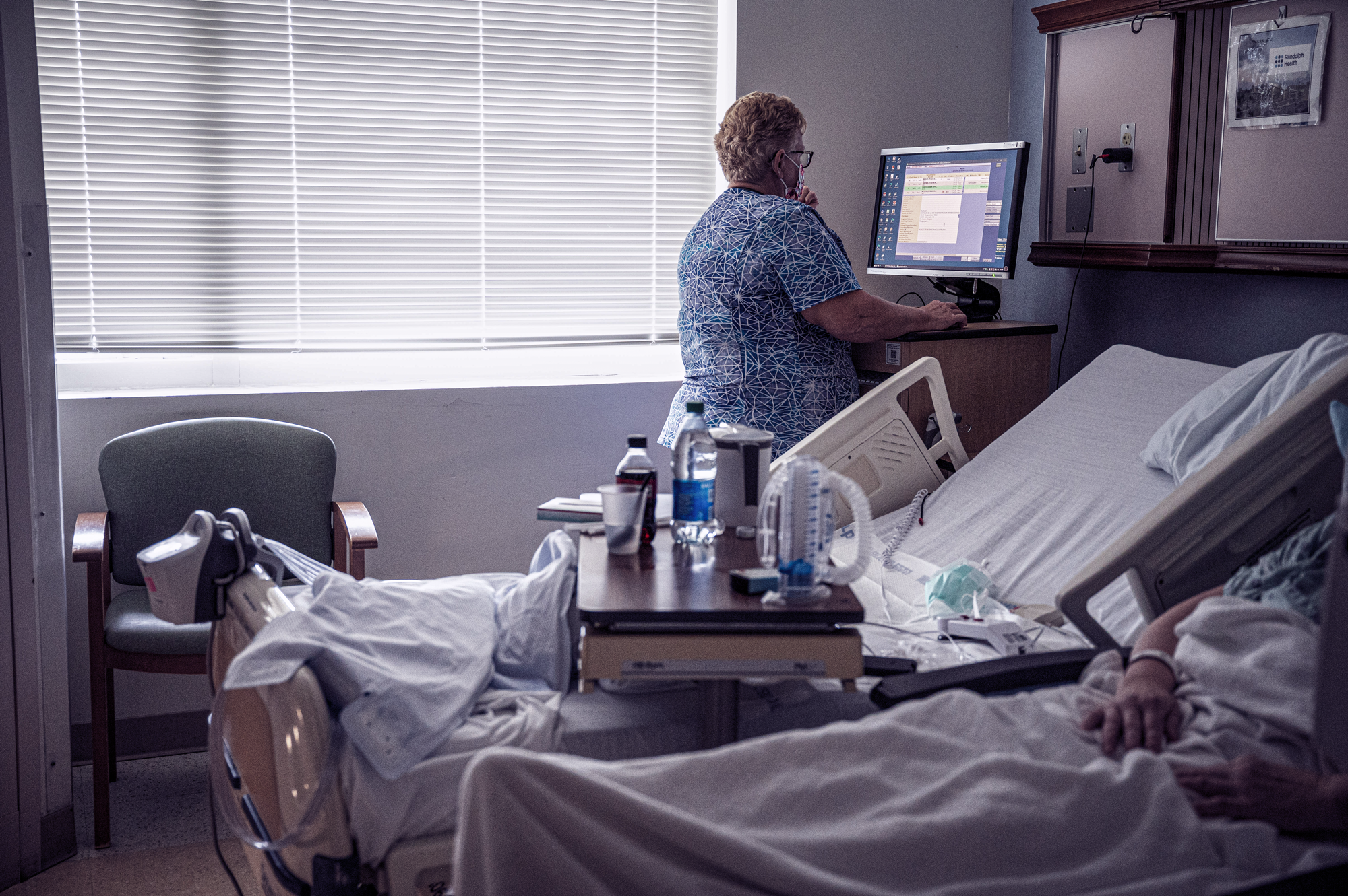A photograph of a nurse seen from behind as she looks at the screen of a computer. Beside her are two hopsital beds, one of which has a patient lying underneath covers, who is visible only from the mid-chest down.