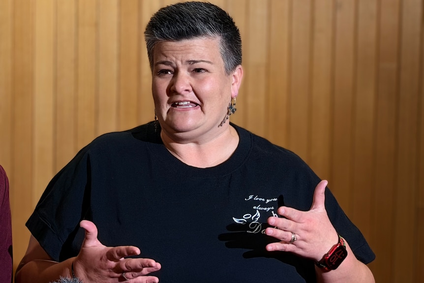 A woman with short hair and a black t-shirt speaks while moving her hands.