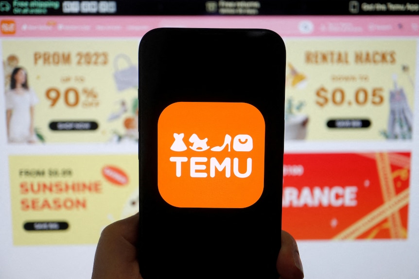 An orange logo with the word TEMU is seen on the screen of a phone held up in front of ads on a computer.