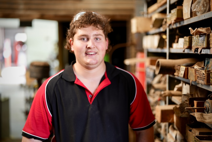 Teenage boy with curly hair smiles at camera wearing black and red mechanic uniform, standing next to busy shelves