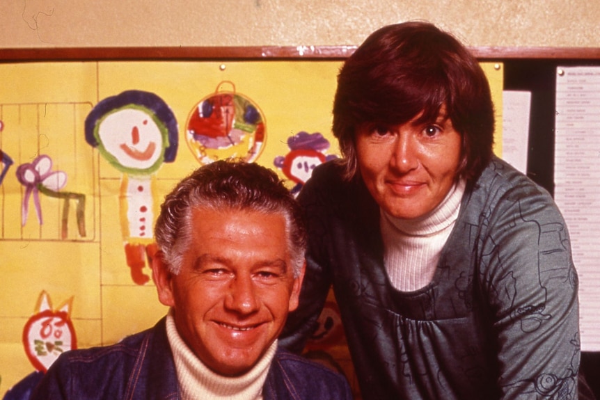A 1960s colour image a man seated at a desk covered in papers, holding a mug. A woman leans over his left shoulder