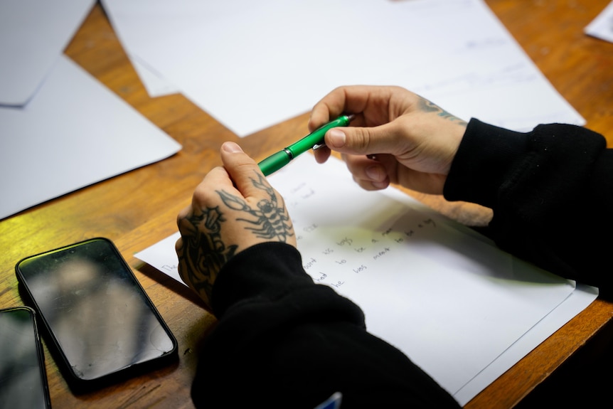 A young man's tattooed hands holding a green pen, resting his hands above a white piece of paper with writing and a phone.