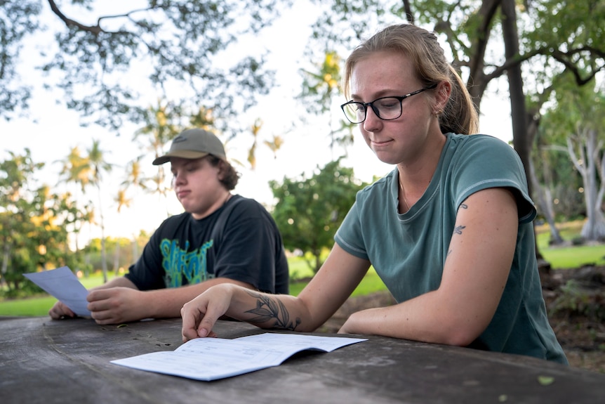 Two people sitting at a table amongst greenery. Young woman in foreground with focussed expression looking down at paper.