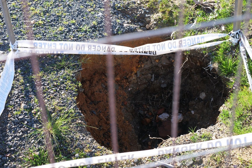 A large hole behind a fence, bitumen and grass around.