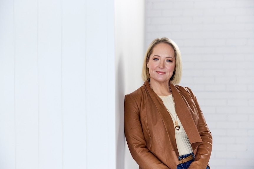 A woman with a blonde bob wears a camel jacket and a cream top while leaning against a white wall.