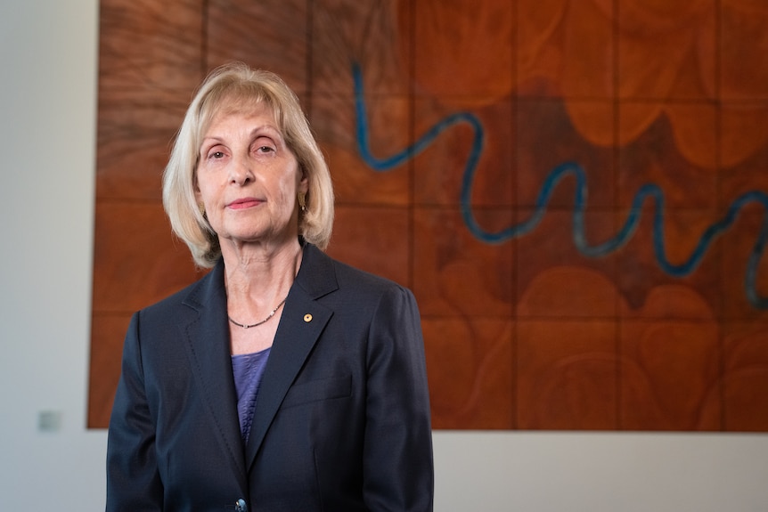 An older woman in a navy-coloured suit in front of an ochre painting.