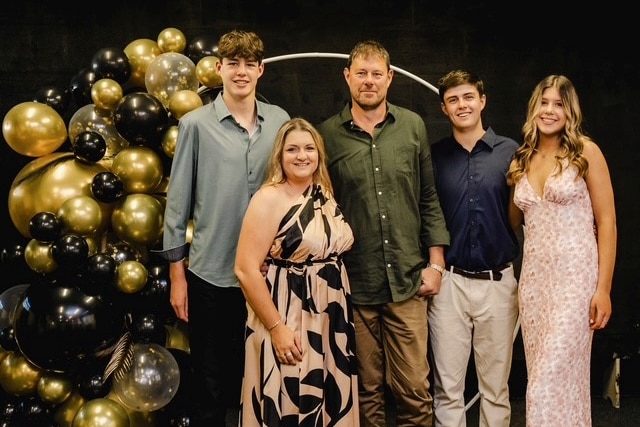 Parents and their three children standing together near a group of black and gold balloons.