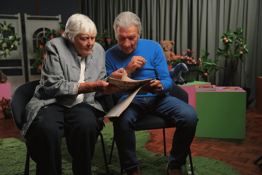 Henrietta and Don, both seated with a colourful set behind them, flick through a book