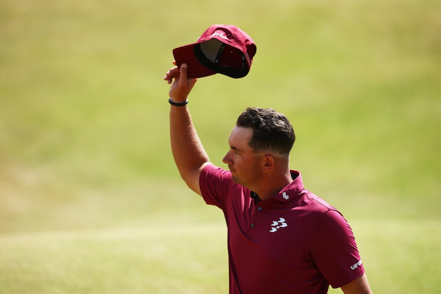 A golfer in a maroon shirt lifts his white cap to acknowledge crowd on fairway