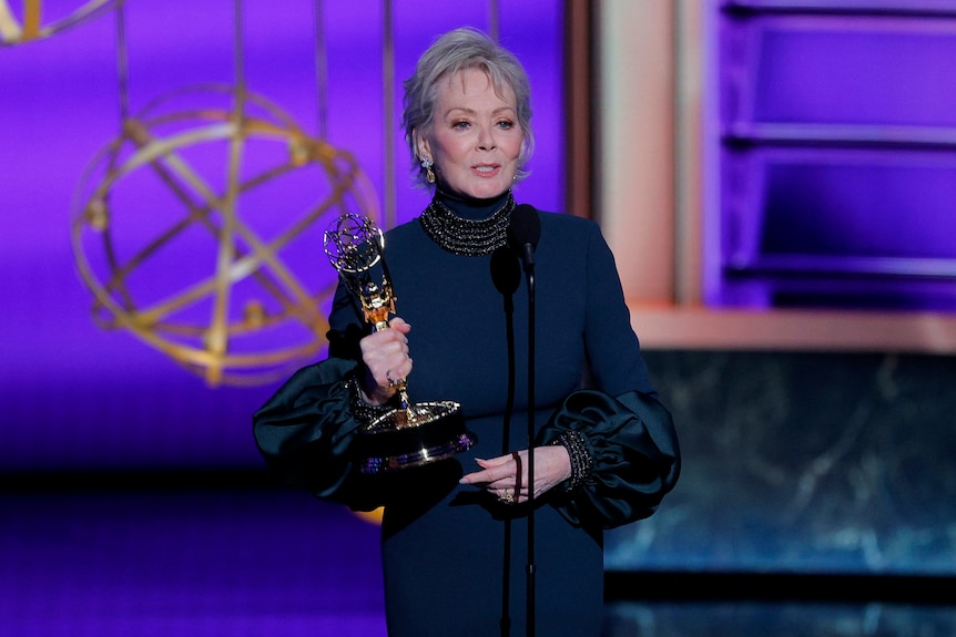 Jean Smart in blue gown holding Emmy statue and talking on stage.