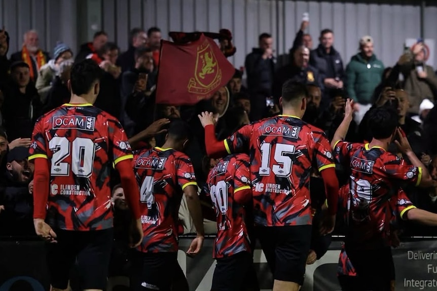 Men in red and black jersey with their back to the camera as people look on. Flags flying.