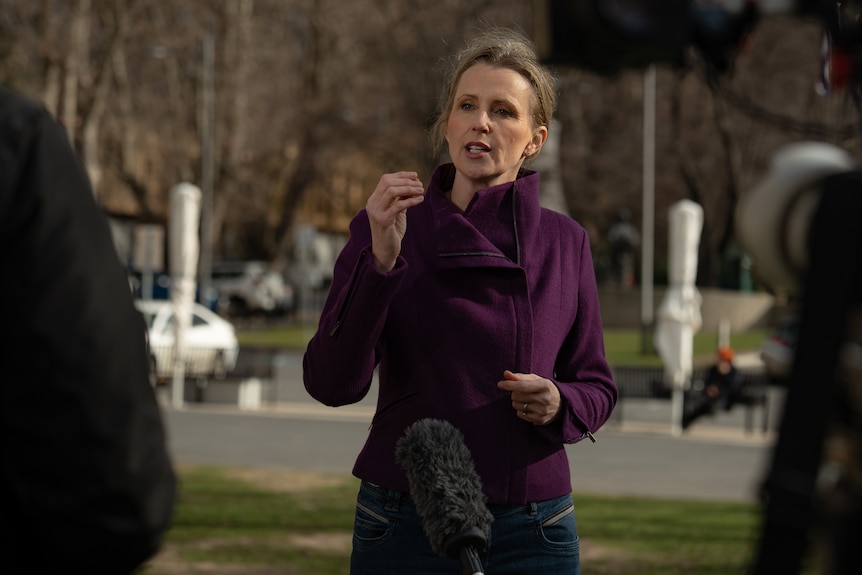 A blonde woman in a maroon blazer talks passionately to a journalist