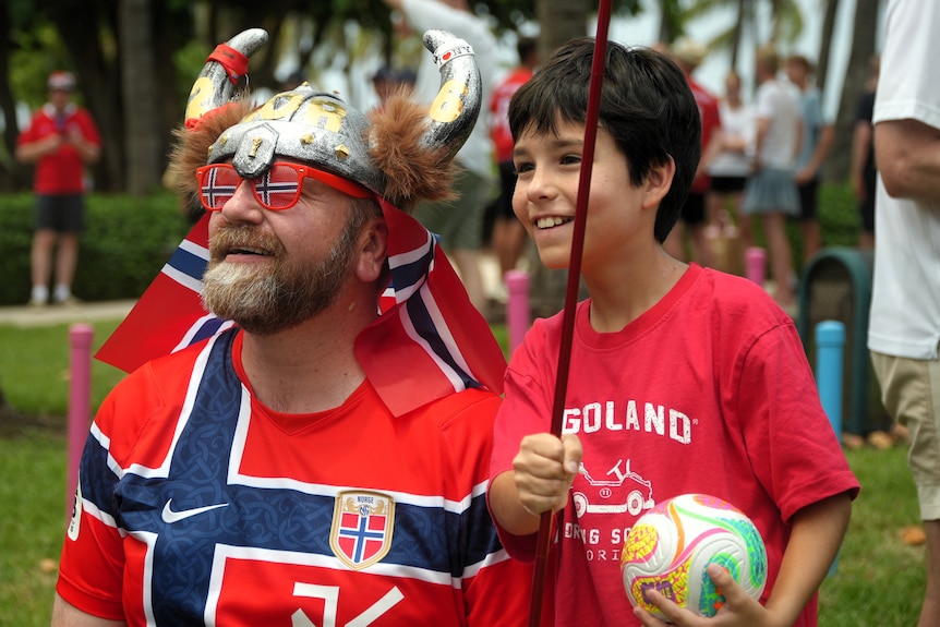 A Norwegian man wearing a Viking helmet, Norway flag glasses, and a jersey, poses with a young boy.