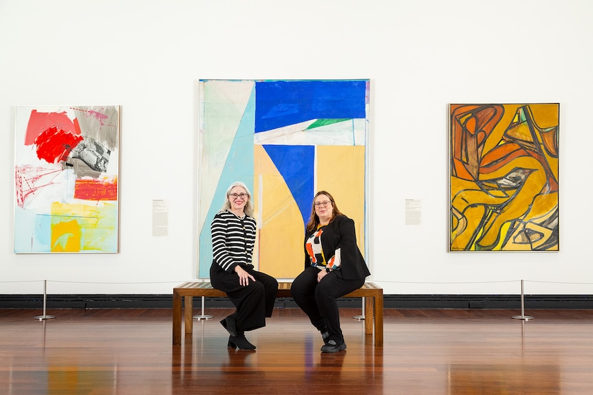 Two women sitting on a wooden bench in front of three paintings hung on a white gallery wall.