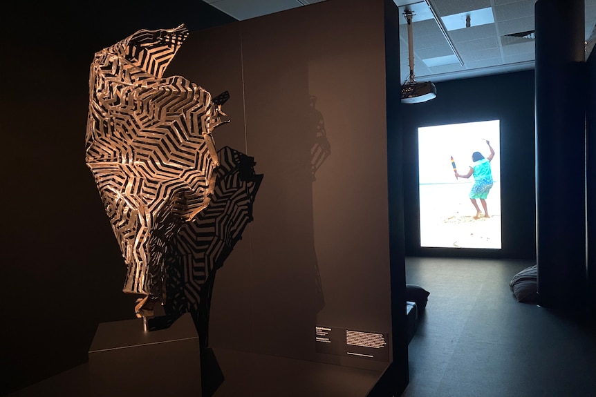 A metal sculpture on display inside an art gallery, in the background a video plays footage of an Indigenous person dancing.