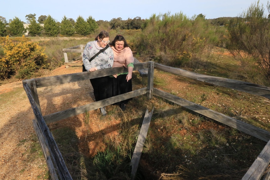 Natasha points down a mineshaft to another woman, scrub, bush surrounds.