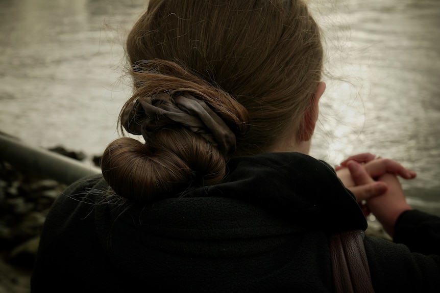 A close of a woman's hair styled in a bun. Her hands are clasped together with a river in the background.