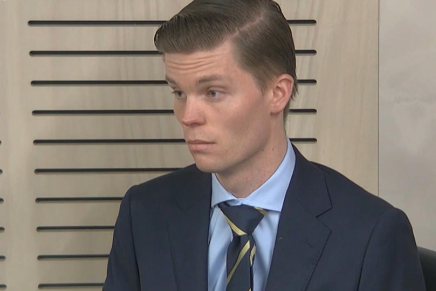 A man in a suit and tie and blue shirt with serious face, wood panelling behind him