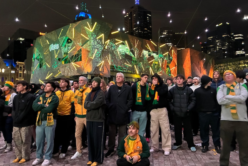 A crowd of people in winter clothing stand and watch big screen with colourfully lit building behind them