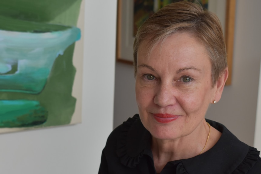 A headshot of woman with short hair and red lipstick, a painting is in the background