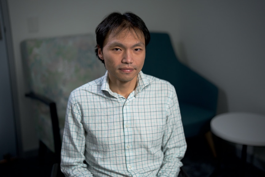 An Asian man in a white shirt with a checked pattern sitting in a chair.