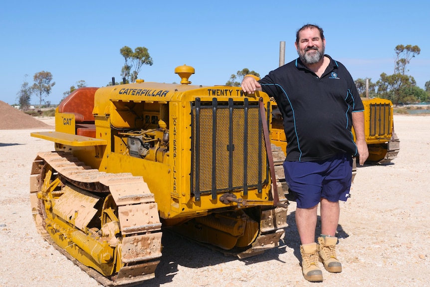 man smiles leaning against small yellow tractor