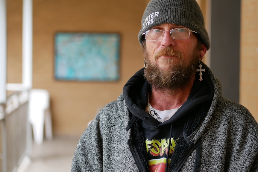 A man in a beanie and a hoodie stands under the awning of a building.