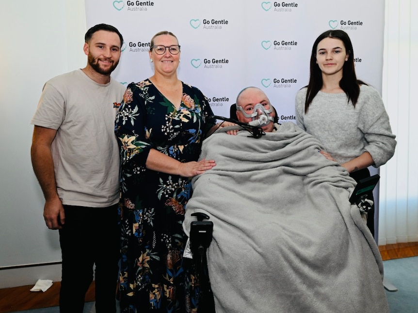 A family of four stand before a Go Gentle Australia banner. Jason a wheelchair user has Motor Neurone Disease.
