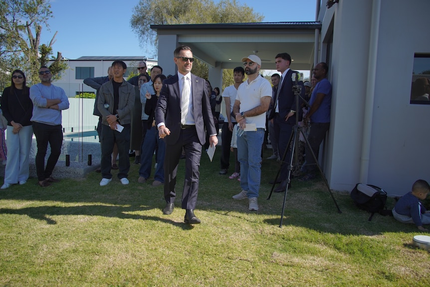 A real-estate agent wearing a suit and sunglasses walks through a crowd of people at an auction.