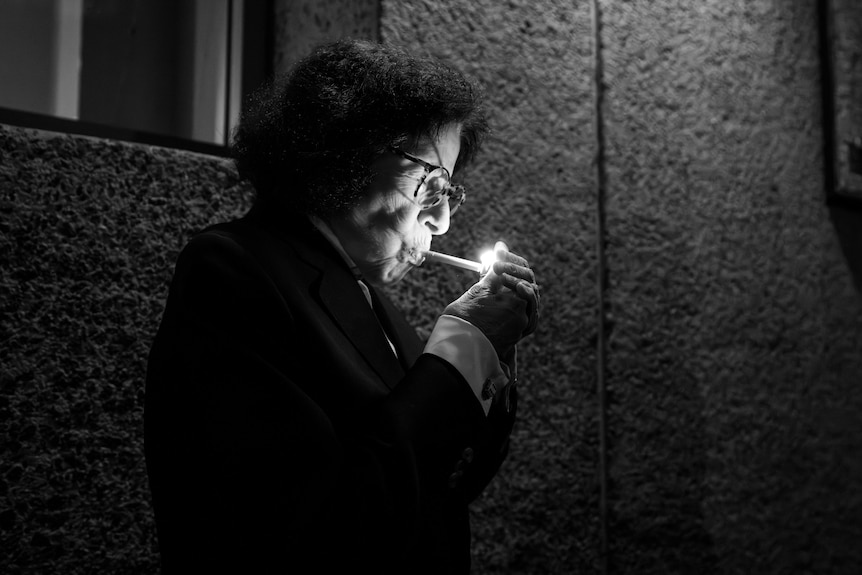 Black and white close up of Fran Lebowitz in profile as she lights up a cigarette, wearing her signature dark jacket white shirt