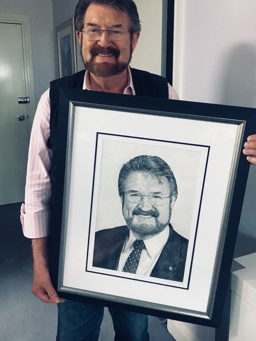 A dark haired, bearded man wearing glasses holds a framed black and white photo of himself.