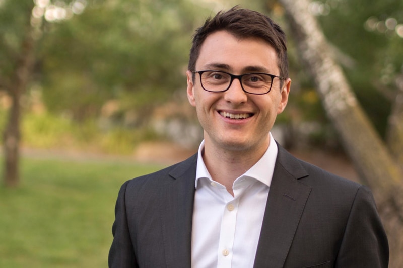 A man with short dark hair wearing a black suit jacket stands outdoors smiling.
