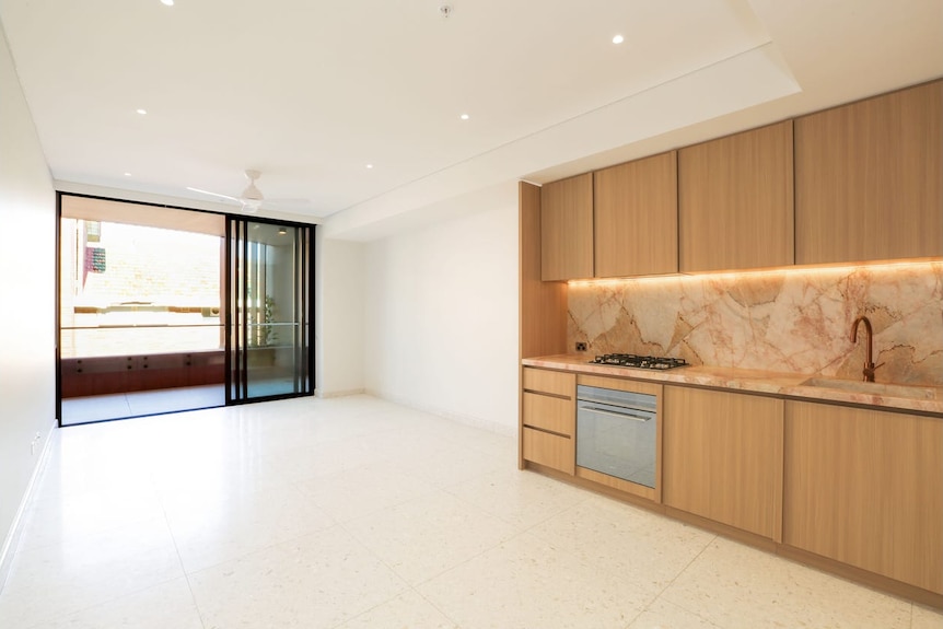 A real estate photo shows a modern galley kitchen opening on to a vacant living area.