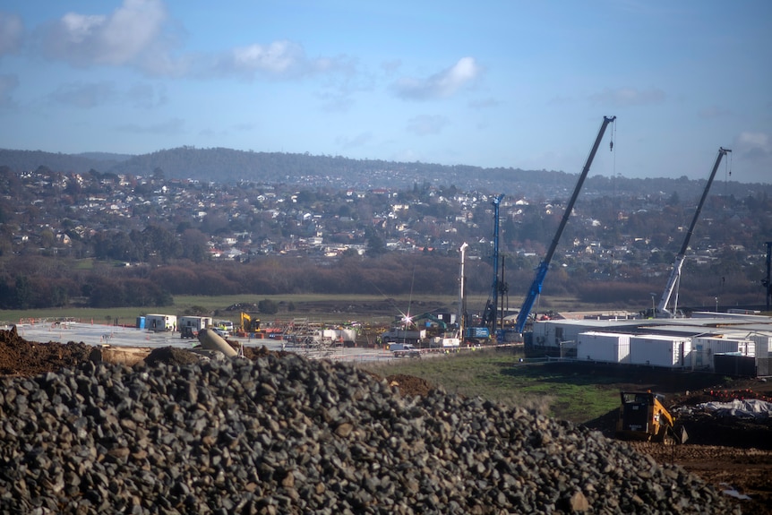 Blue cranes at a distant construction site in a valley dotted with homes under a blue hazy sky.