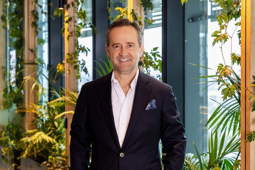 A dark-haired man in a blazer stands in an office with a number of plants near the windows.