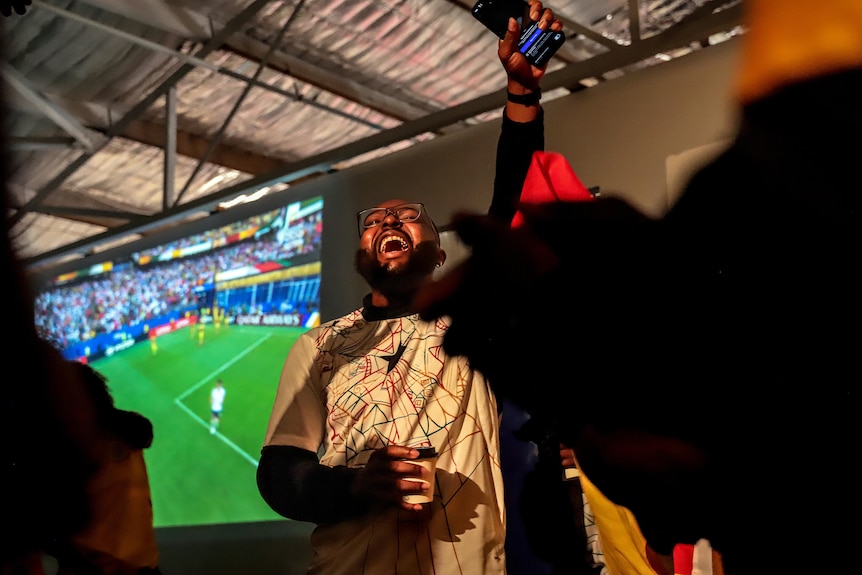 A man stands in front of a screen with his hand in the air. There's people around him and football showing on the screen behind.