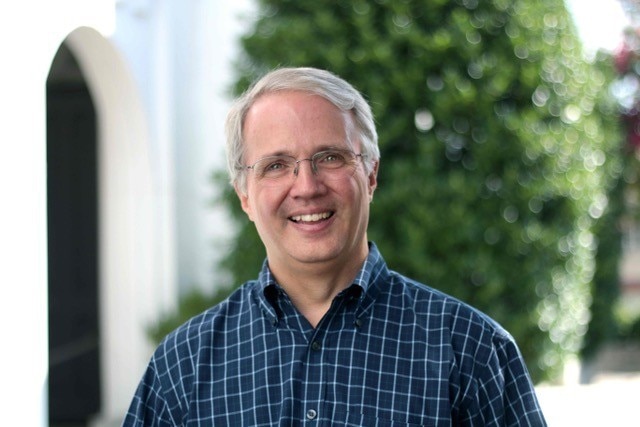 Smiling man with grey, cropped hair and glasses, blue button down checked shirt, headshot outside.