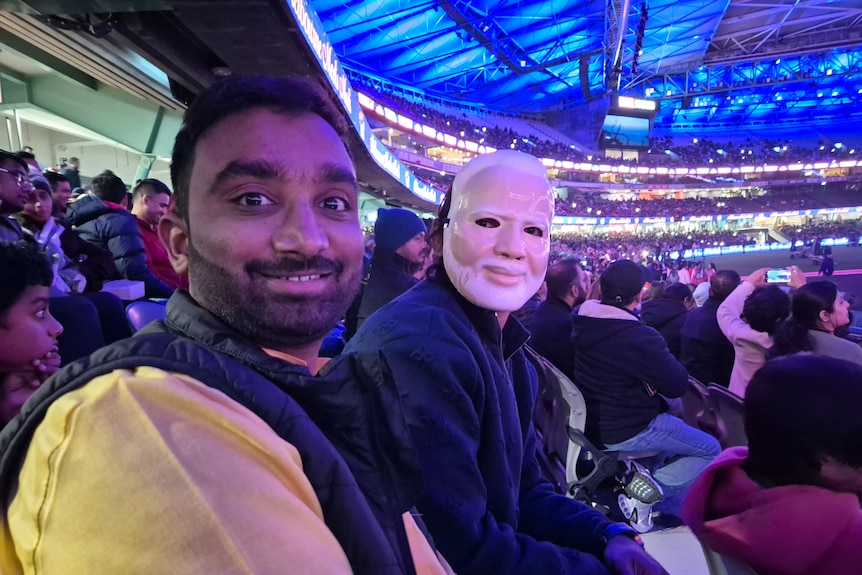 Two men smile at the camera while sitting in a big stadium, one wears a mask.