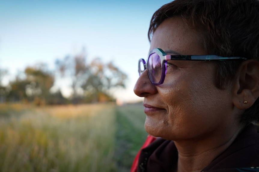 A close-up side profile of a woman wearing a neutral facial expression, looking over the vast outback landscape before her.