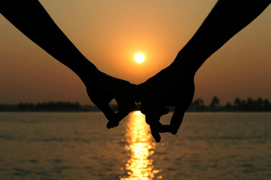 Photo of two people's hands with their fingers entwined, in silhouette, in front of a river