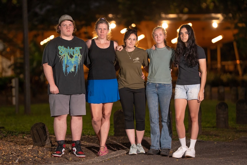 One young man and four women standing together at night, looking at camera with a serious expression, arms around each other.