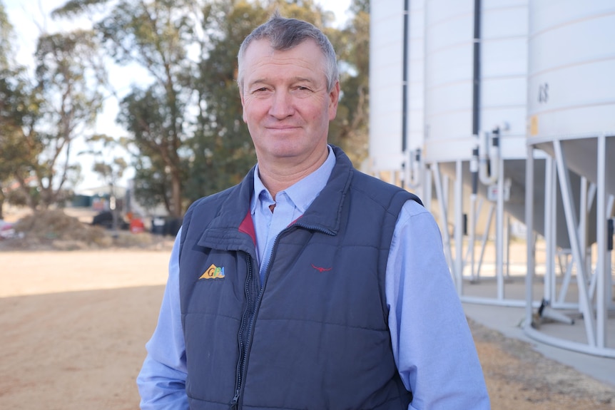 man with short grey and white hair, wearing denim shirt and blue padded vest stands in front of 3 white grain silos